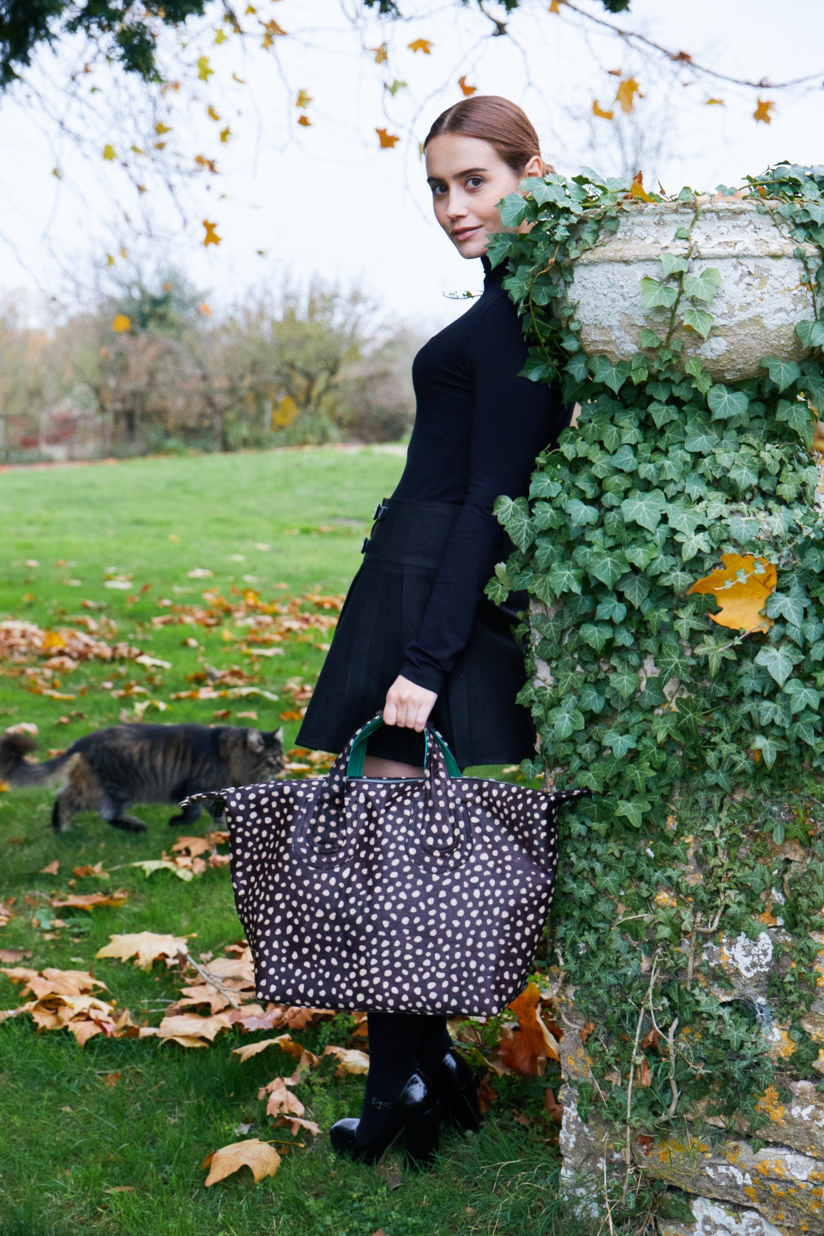 Woman in a black dress stands beside an ivy-covered stone column, holding a large polka-dot Brix + Bailey tote bag. Autumn leaves and a curious cat complete the countryside charm.