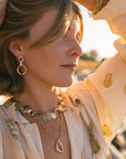 Woman wearing handcrafted Rebecca J Designs jewelry in golden sunset light - layered necklaces and circular earrings showcasing artisanal metalwork and organic forms"