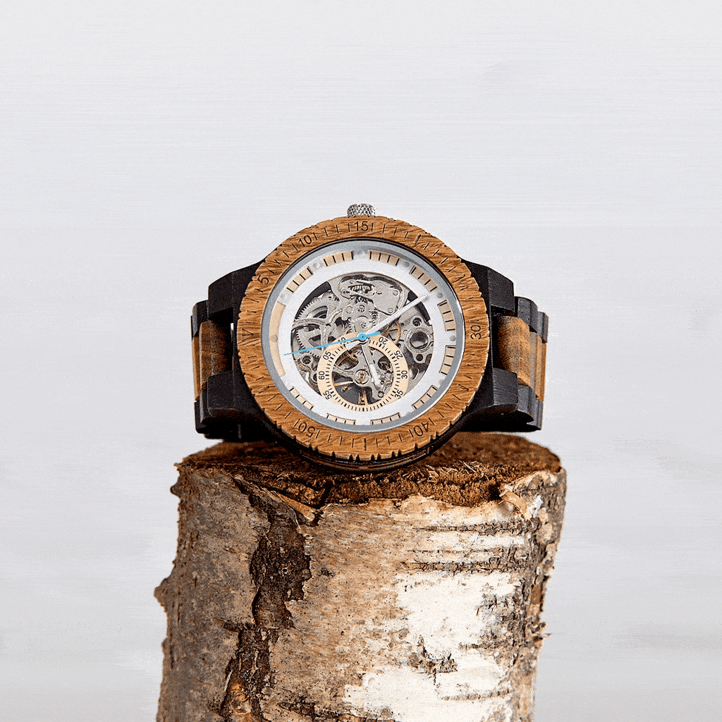 Eco-conscious wooden wristwatch with visible clockwork detail, photographed on a tree trunk with a hummingbird in flight — part of the Brix + Bailey Ethical Brands collection.