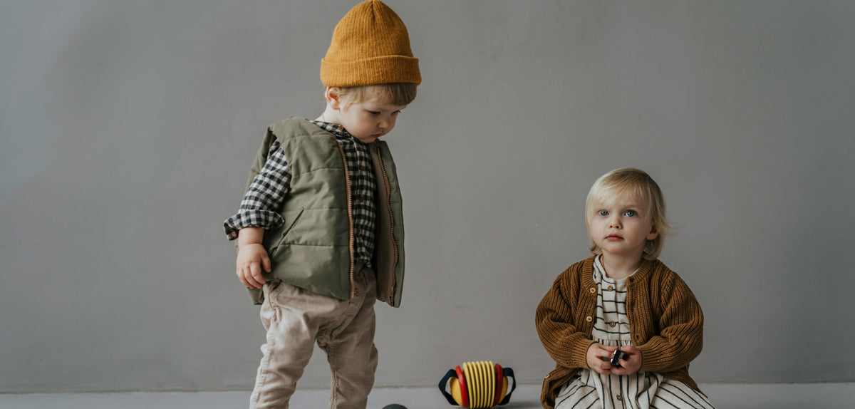 Two young children in earthy handmade knitwear and a mustard beanie, one standing and one sitting with a wooden toy