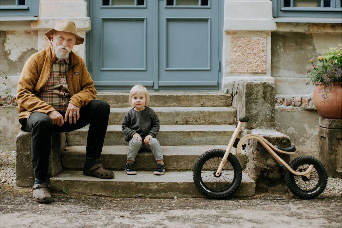 A man in a hat sitting on stone steps beside a small child and a wooden balance bike outside a blue-doored shopfront