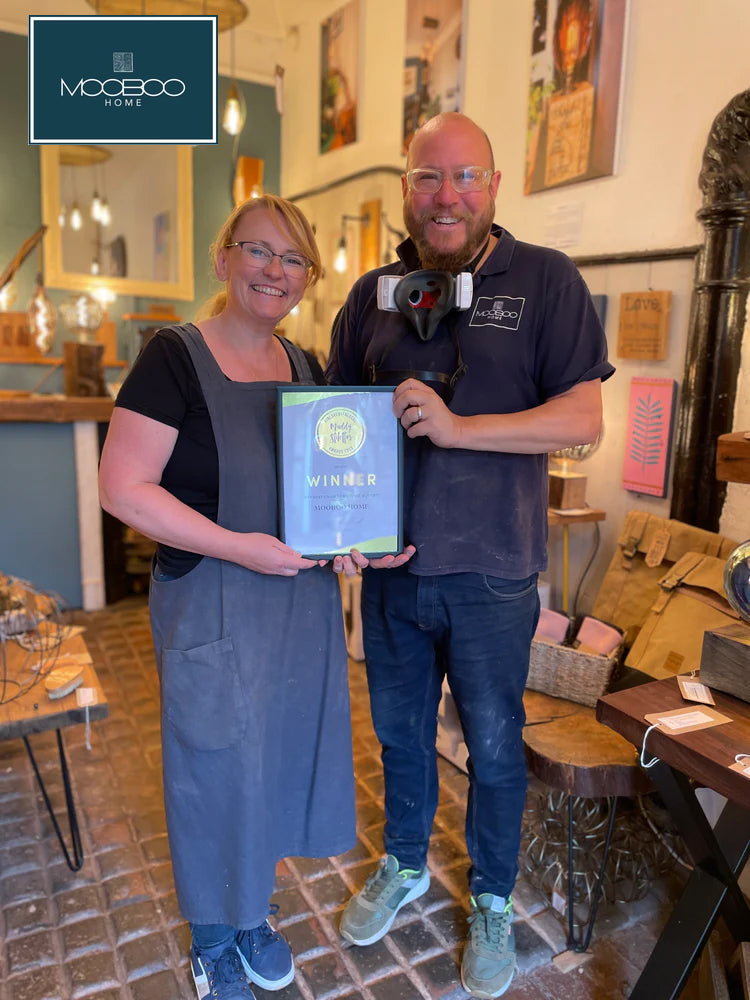 The founders of MooBoo Home in their shop, holding a framed award, with furniture and pictures behind them