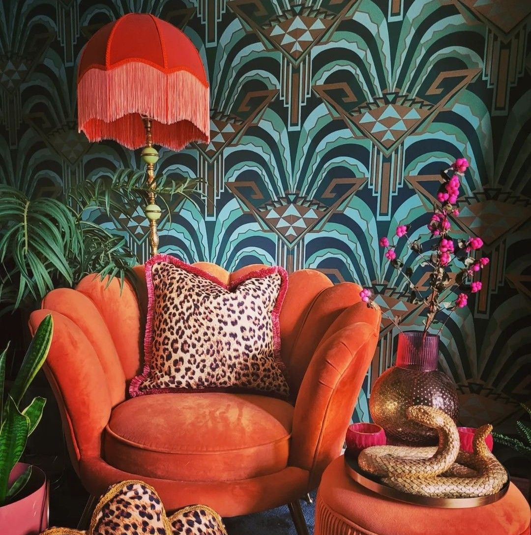 Art deco fan-pattern wallpaper in teal and gold behind a burnt orange velvet armchair with leopard print cushion and fringed lamp
