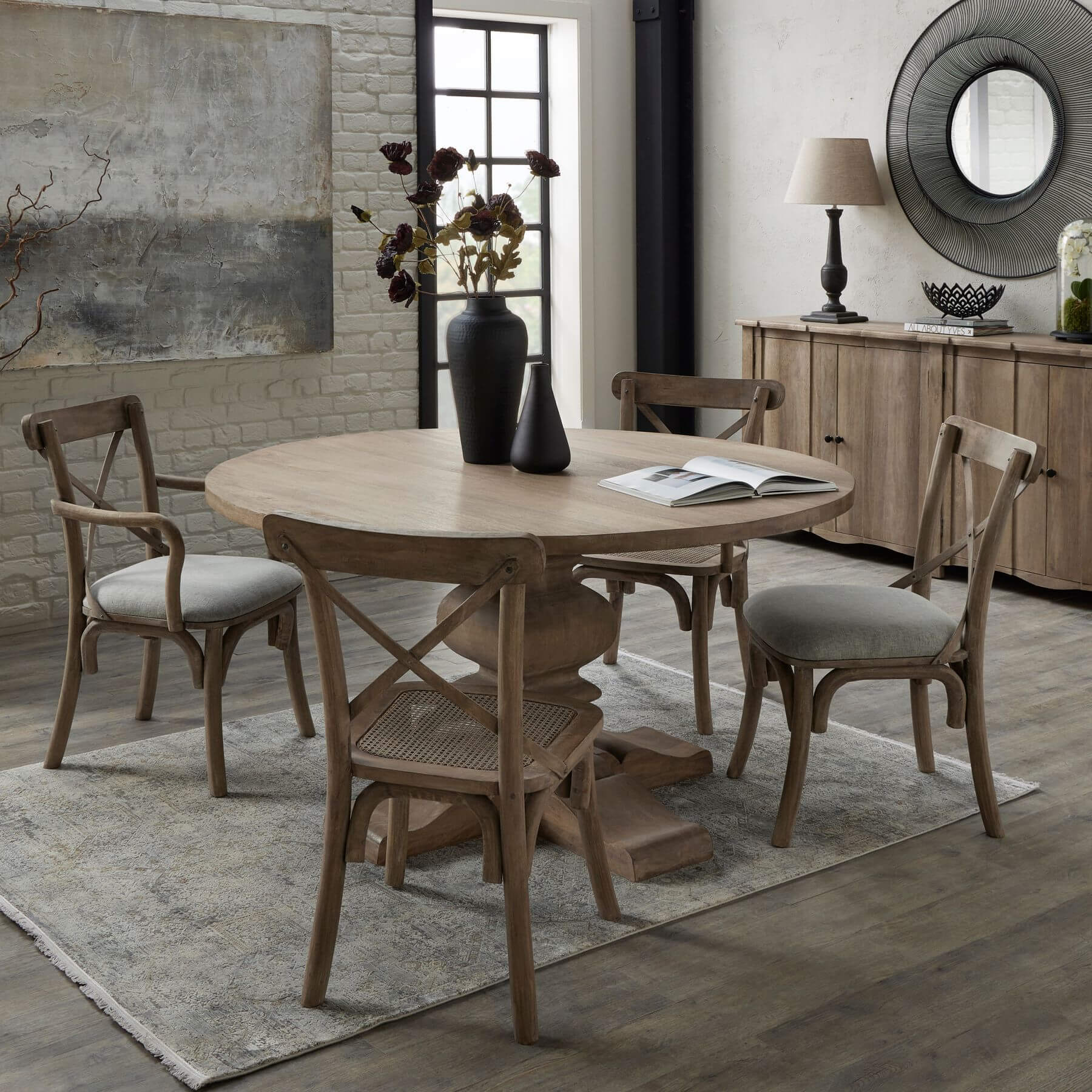 Round hardwood dining table with bleached finish, black iron pedestal base, and warm brown tones for elegant gatherings. by Sostter at brixbailey.com