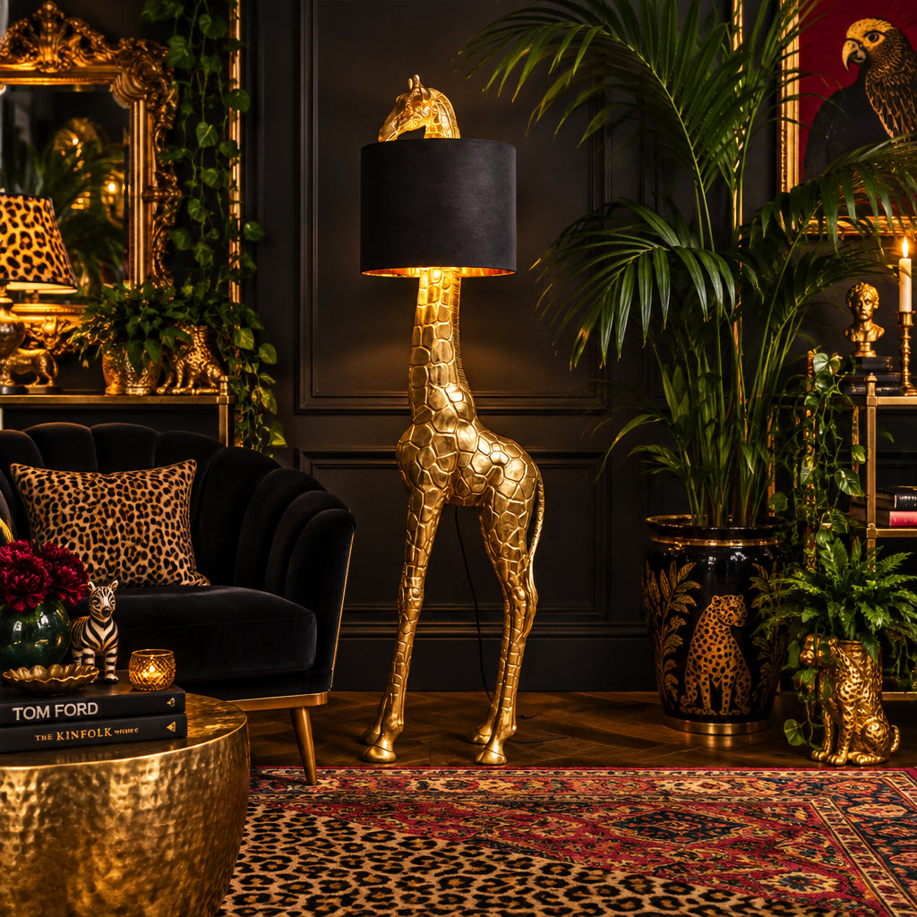 A giraffe-shaped floor lamp with a textured gold finish and a black cylindrical shade, shown lit against a neutral gray studio background.
