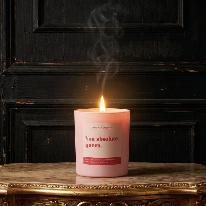 Mischief Candles pink candle labelled "You absolute queen", lit on a gilded marble-topped table against dark wood panelling