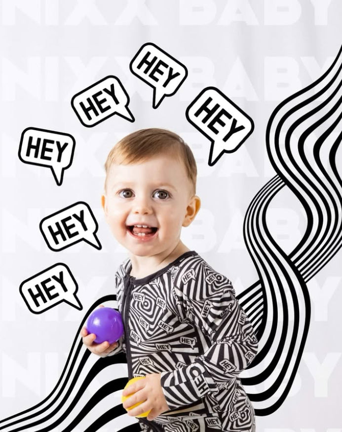Smiling toddler in a bold monochrome Brix + Bailey patterned outfit holding a purple toy ball, surrounded by playful “HEY” speech bubbles and wavy black-and-white graphics — fun, expressive kidswear with attitude.