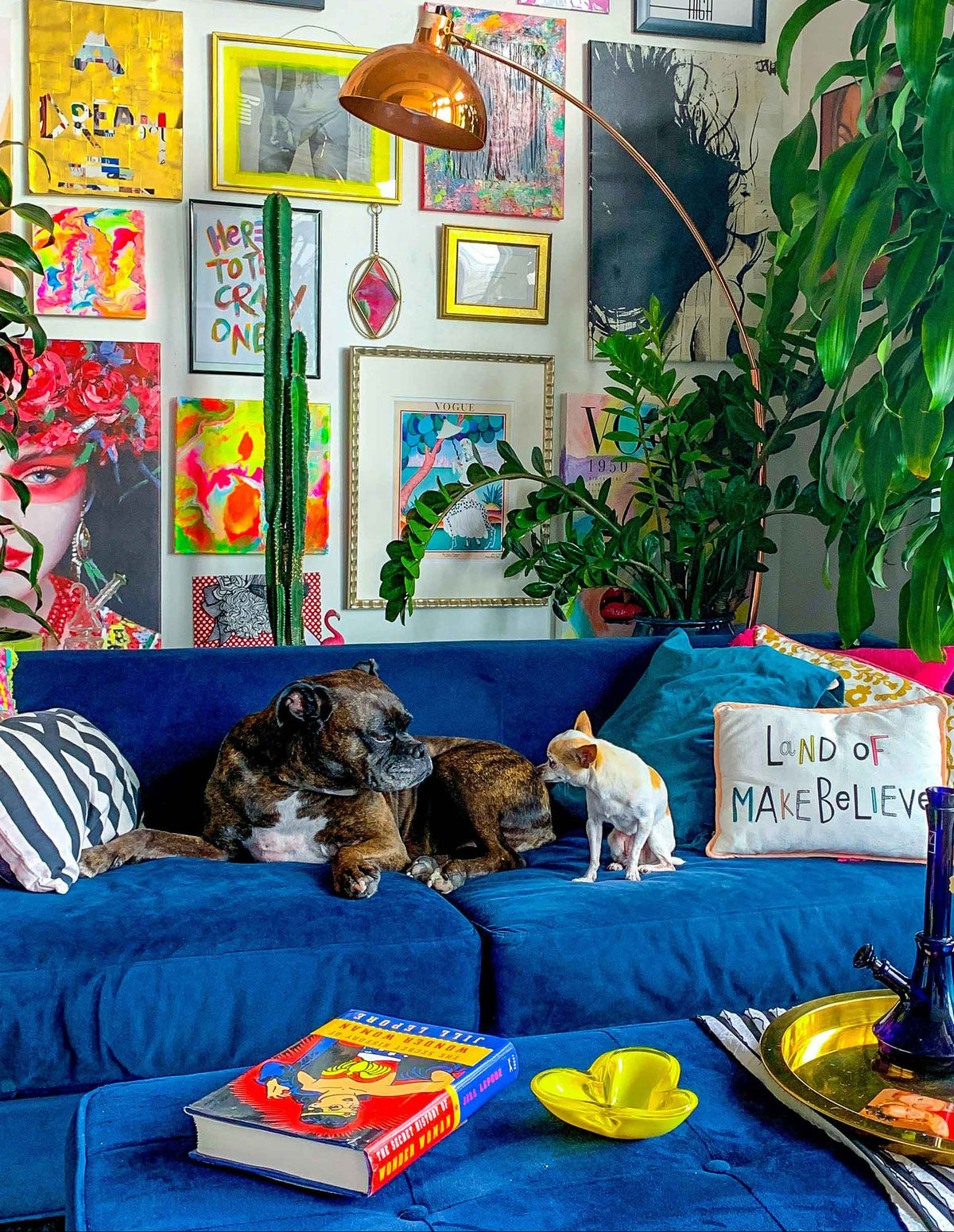 A cozy living room scene with a blue sofa in the foreground, a brachycephalic dog lying down, and a small cat standing nearby. The background is a gallery wall filled with colorful framed art prints, plants, and a copper floor lamp.