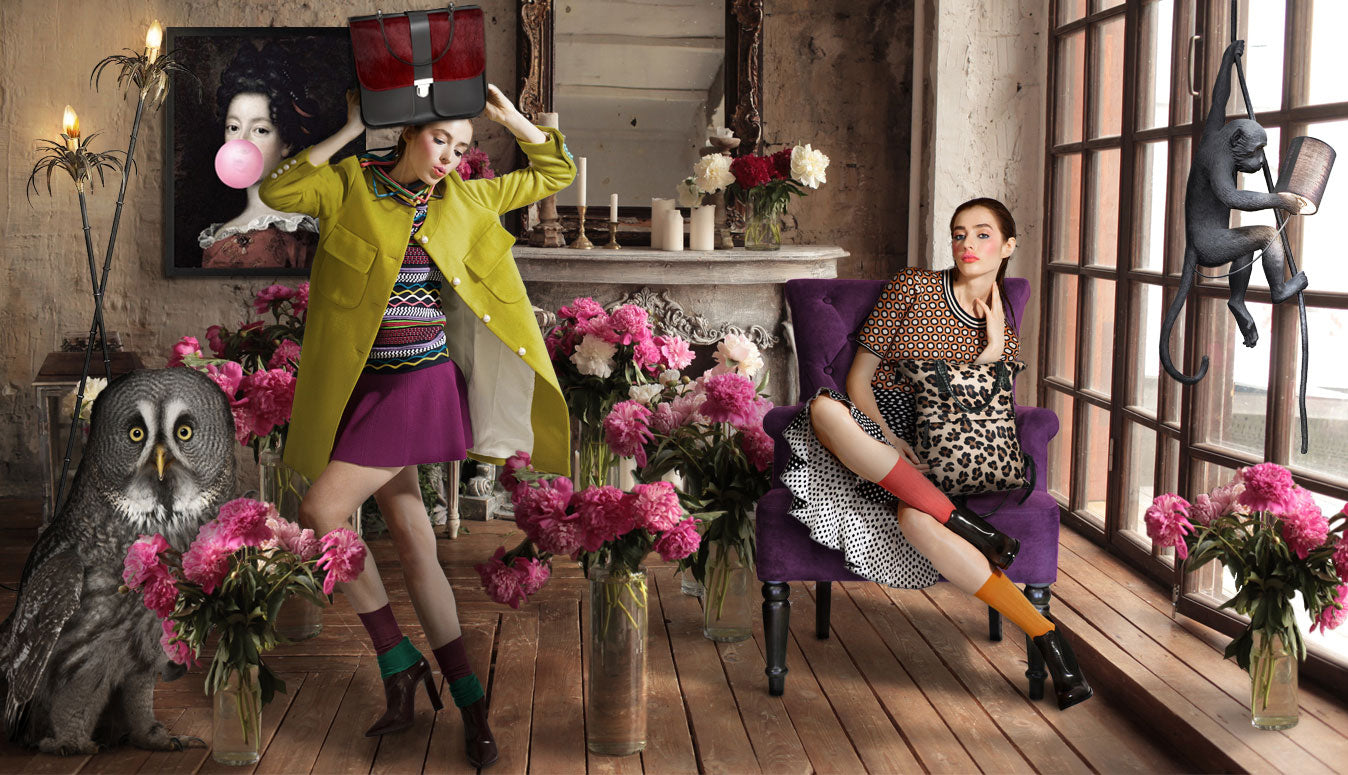 Two stylish women in a curated living space showcasing independent fashion and artisanal home décor - one wearing a chartreuse coat blowing a pink bubble, the other in patterned dress seated on purple velvet chair, surrounded by pink peonies, vintage furnishings, and handcrafted accessories from Brix and Bailey