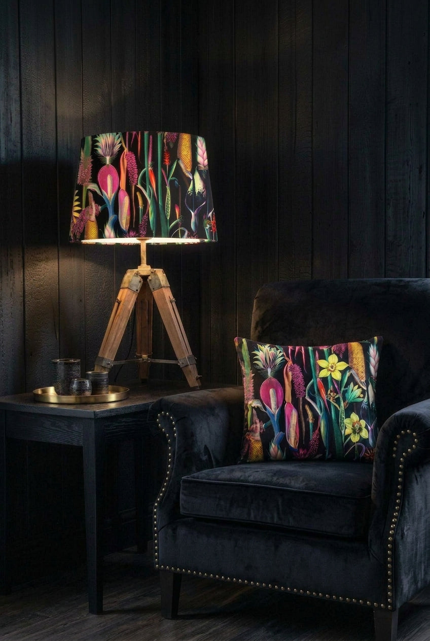 By Poppy handmade lampshade in a bold botanical print on black, lit on a wooden tripod lamp beside a matching cushion