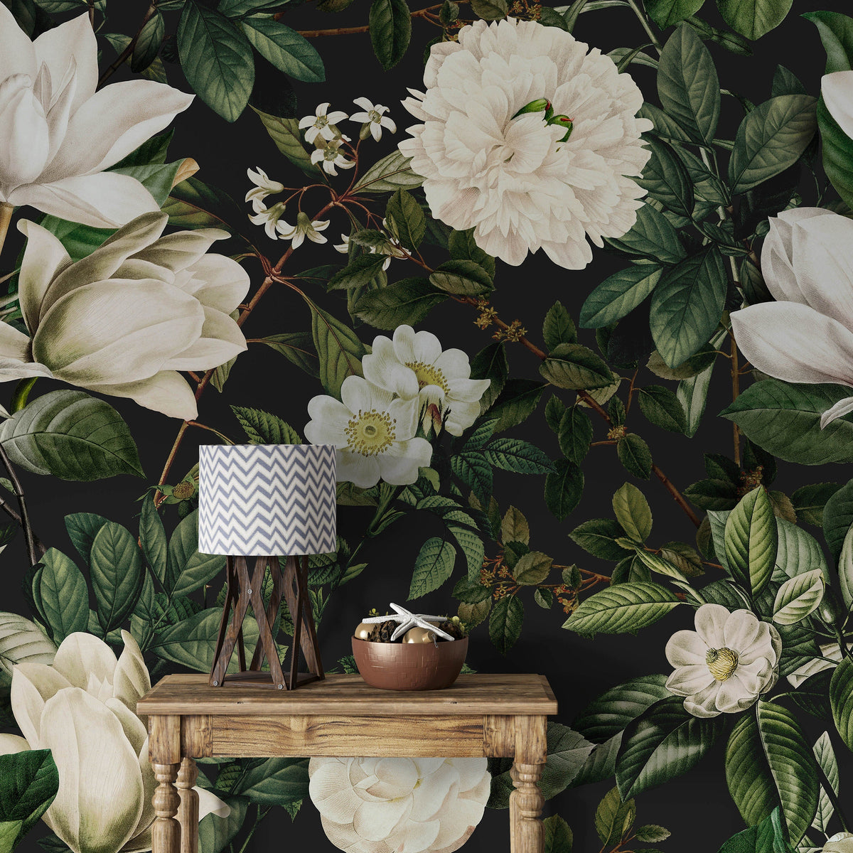 A styled interior scene featuring a dark floral wallpaper with large cream magnolia and white blossoms, green leaves, and a small wooden table holding a lamp and a bowl at Brix + Bailey