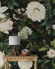 A styled interior scene featuring a dark floral wallpaper with large cream magnolia and white blossoms, green leaves, and a small wooden table holding a lamp and a bowl at Brix + Bailey