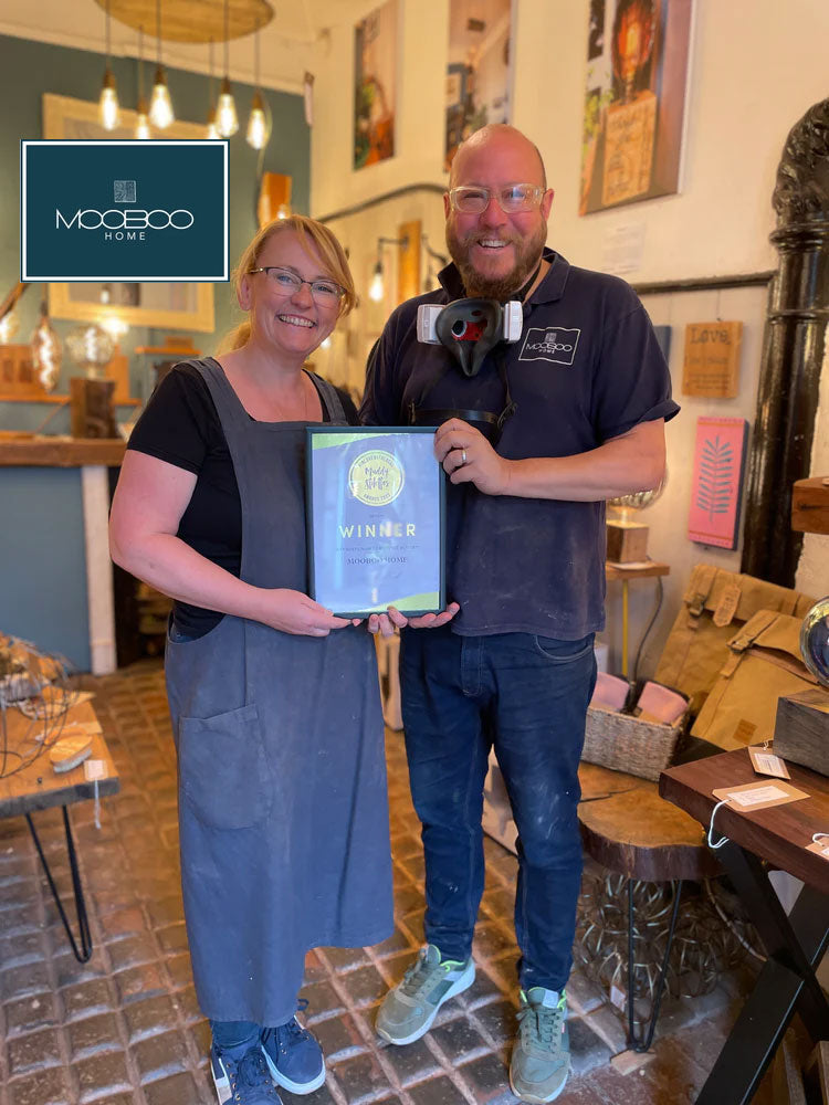 MooBoo Home founders Sarah and Mark in their workshop, holding their sustainability award, surrounded by handcrafted wooden lighting fixtures and workshop tools