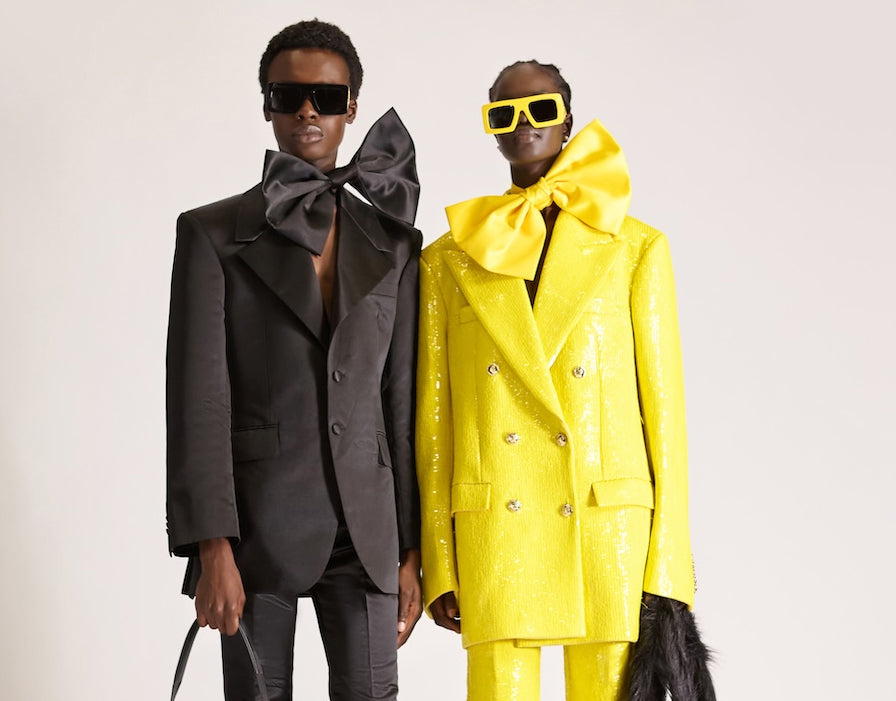 Two models in bold tailored outfits, one in a black suit with an oversized bow and one in a bright yellow double breasted suit with matching bow and sunglasses, photographed against a minimal backdrop for the womenswear curated collection on the Brix and Bailey marketplace. at Brix and Bailey